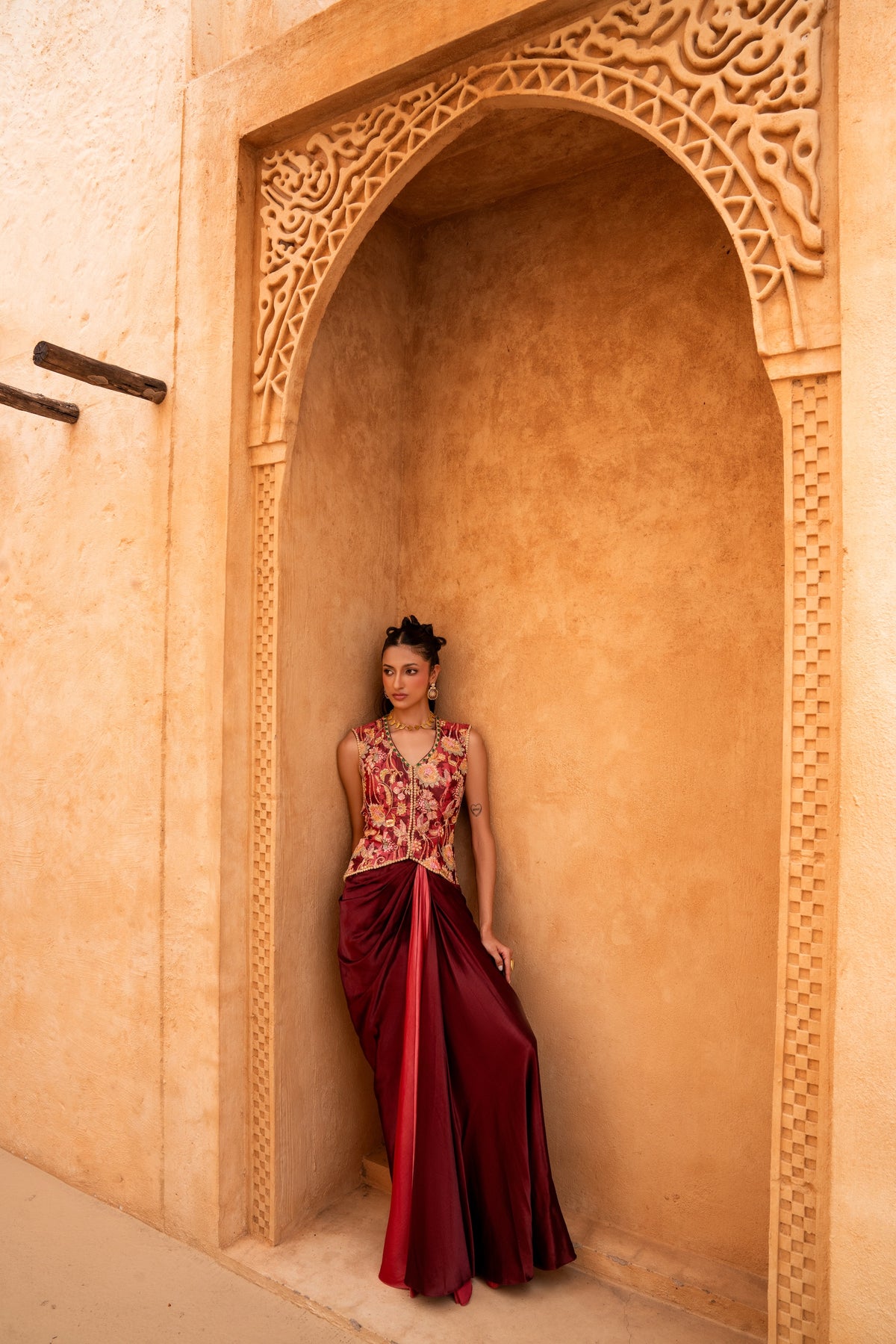 A woman wearing a sleeveless floral-print top and a deep burgundy satin skirt stands inside a carved ochre archway.