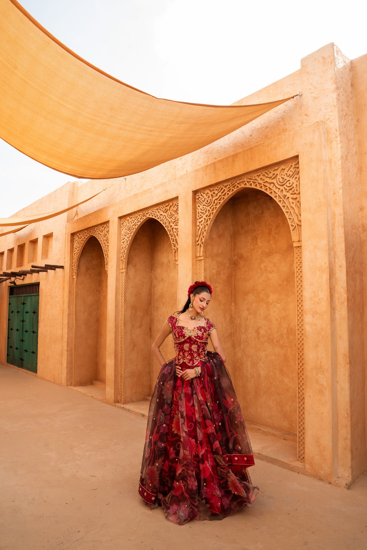 A woman wearing a red and black floral ball gown stands in front of a sandy desert architectural wall with arches and a large beige canopy overhead.