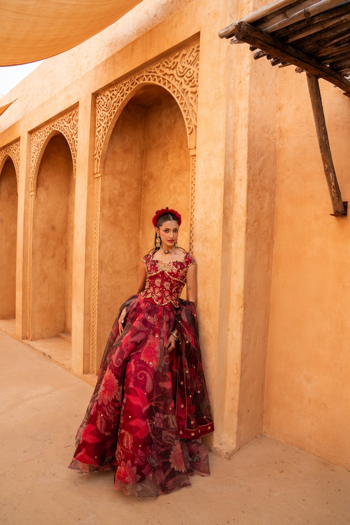 A woman stands in a desert courtyard wearing an ornate red and maroon ball gown with gold embroidery and a matching headpiece, posed in front of tan arches.