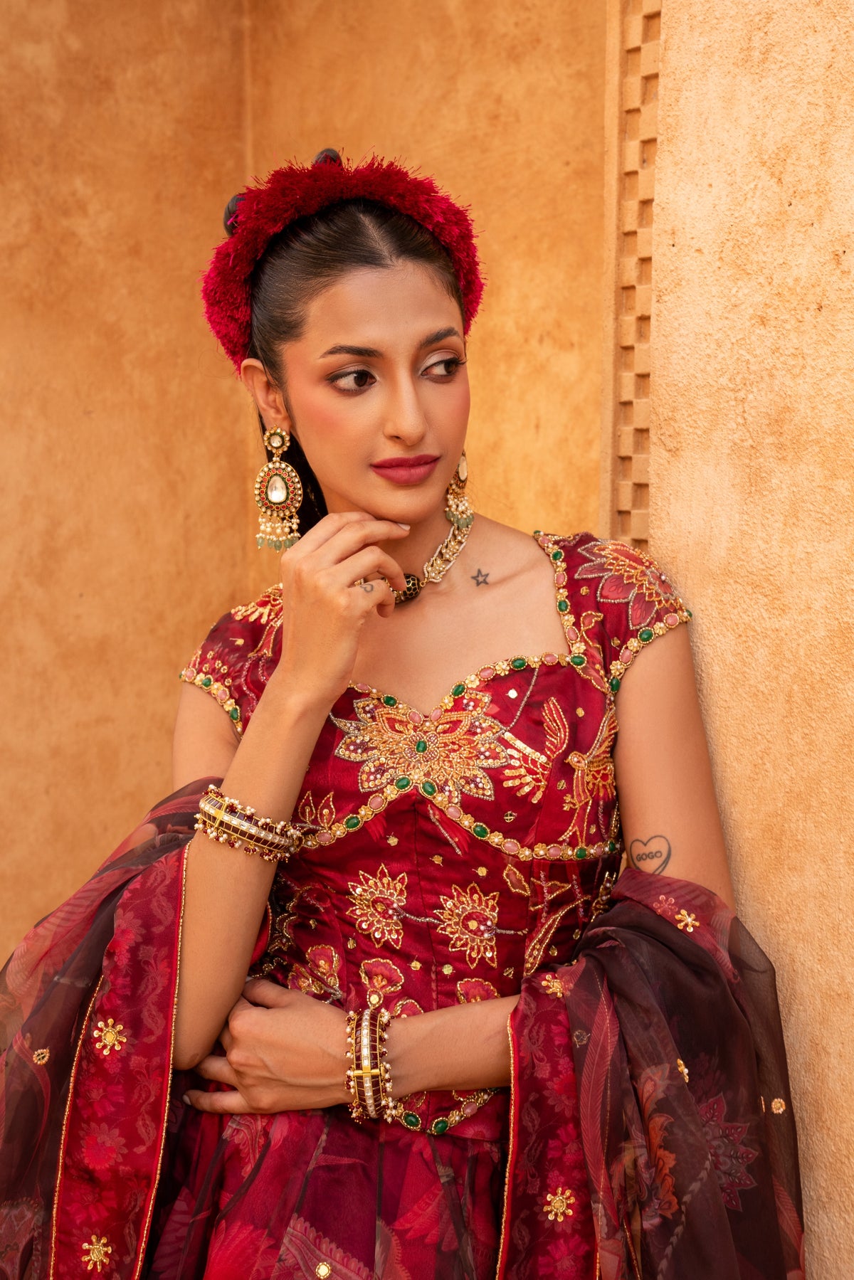 A woman wearing a traditional Indian red and gold embroidered lehenga choli with a matching dupatta, gold jewelry, and a red floral headpiece, posing against a textured beige wall.