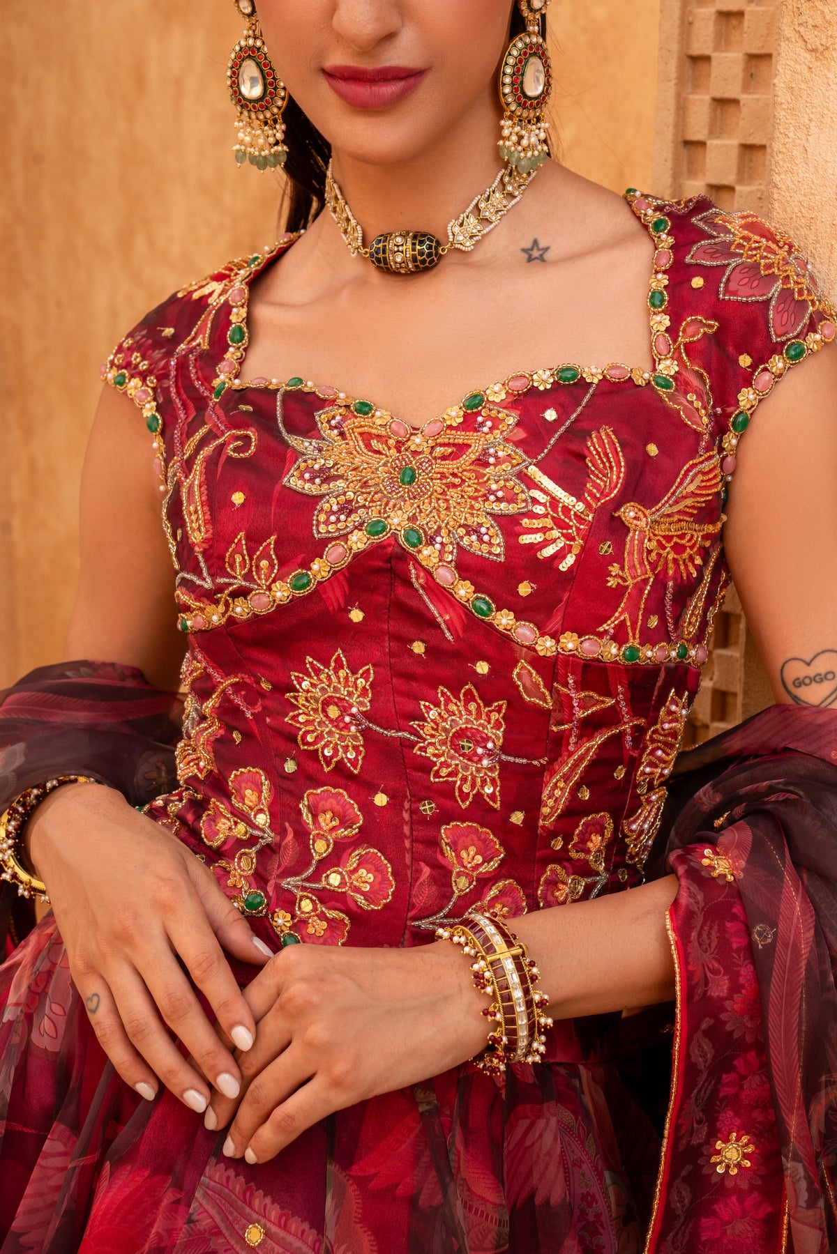 A close-up of a model wearing a rich red and gold heavily embroidered traditional Indian dress with green stone accents, gold jewelry, and a matching sheer dupatta.