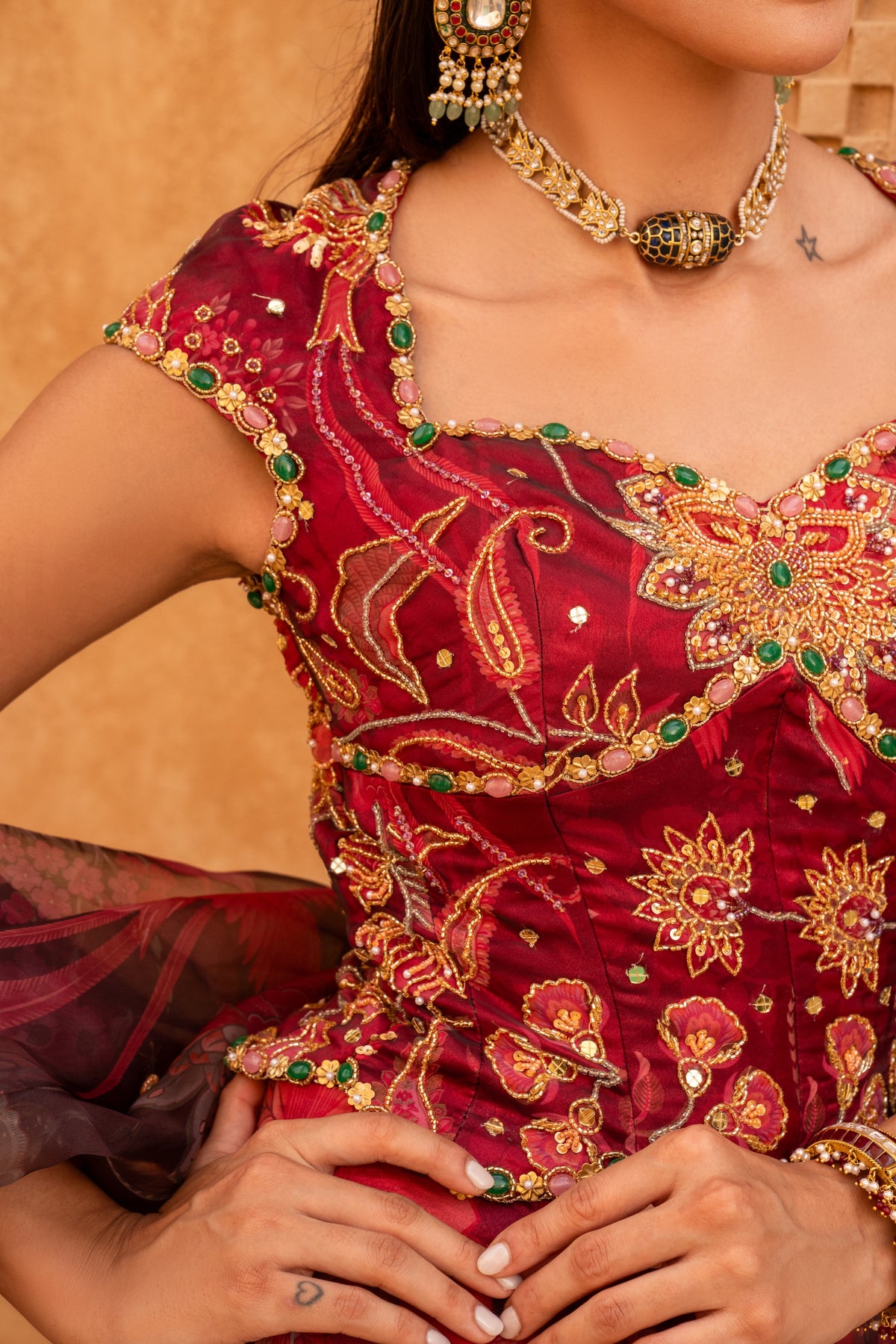 A close-up of a woman wearing an ornate deep red embroidered traditional outfit with gold floral detailing and green jewel accents, paired with matching gold jewelry against a warm beige background.