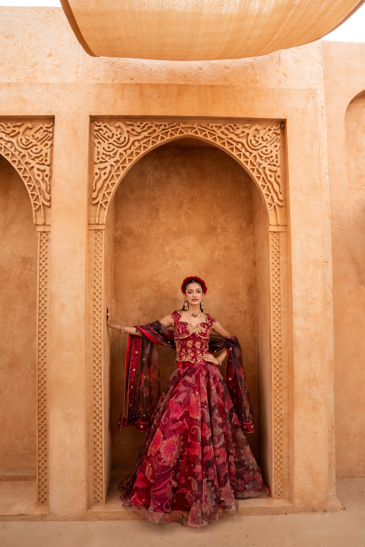 A woman wearing an ornate red and magenta floral lehenga choli traditional Indian gown stands in a carved beige stone archway.