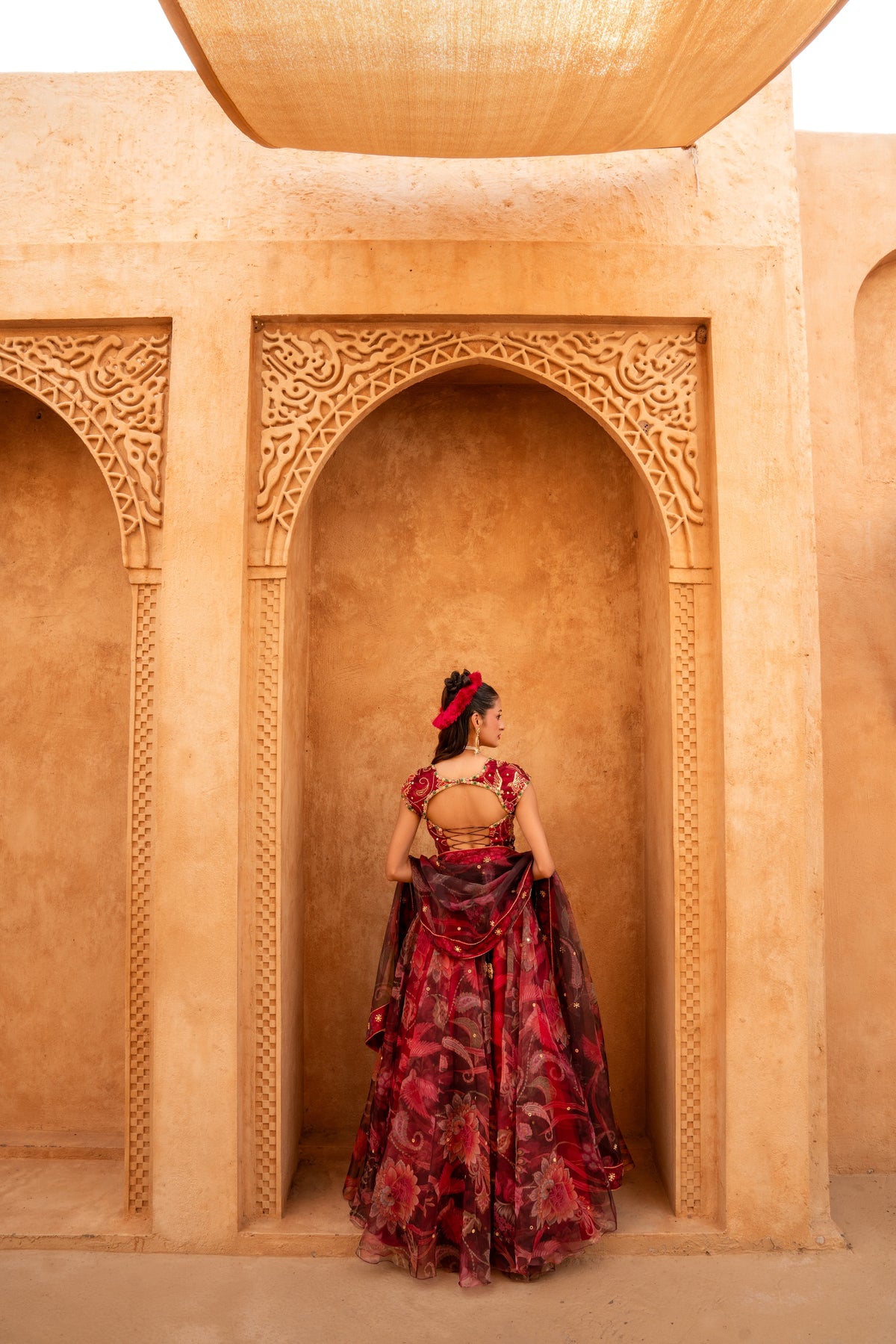 A woman stands with her back to the camera in a desert architectural setting, wearing a deep red and black floral lehenga choli with a sheer dupatta.