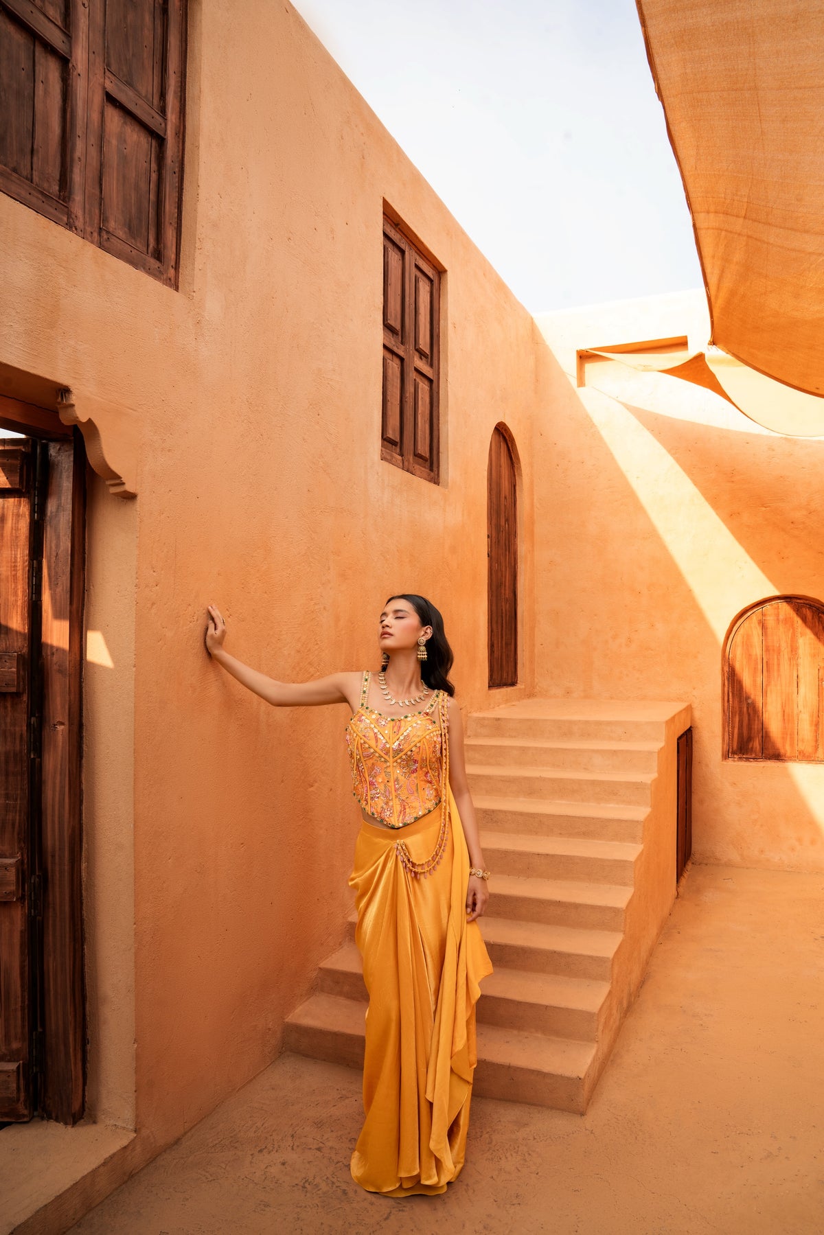 A woman wearing a bright yellow and gold embellished traditional dress stands in a sandy courtyard with terracotta-colored buildings and wooden doors.