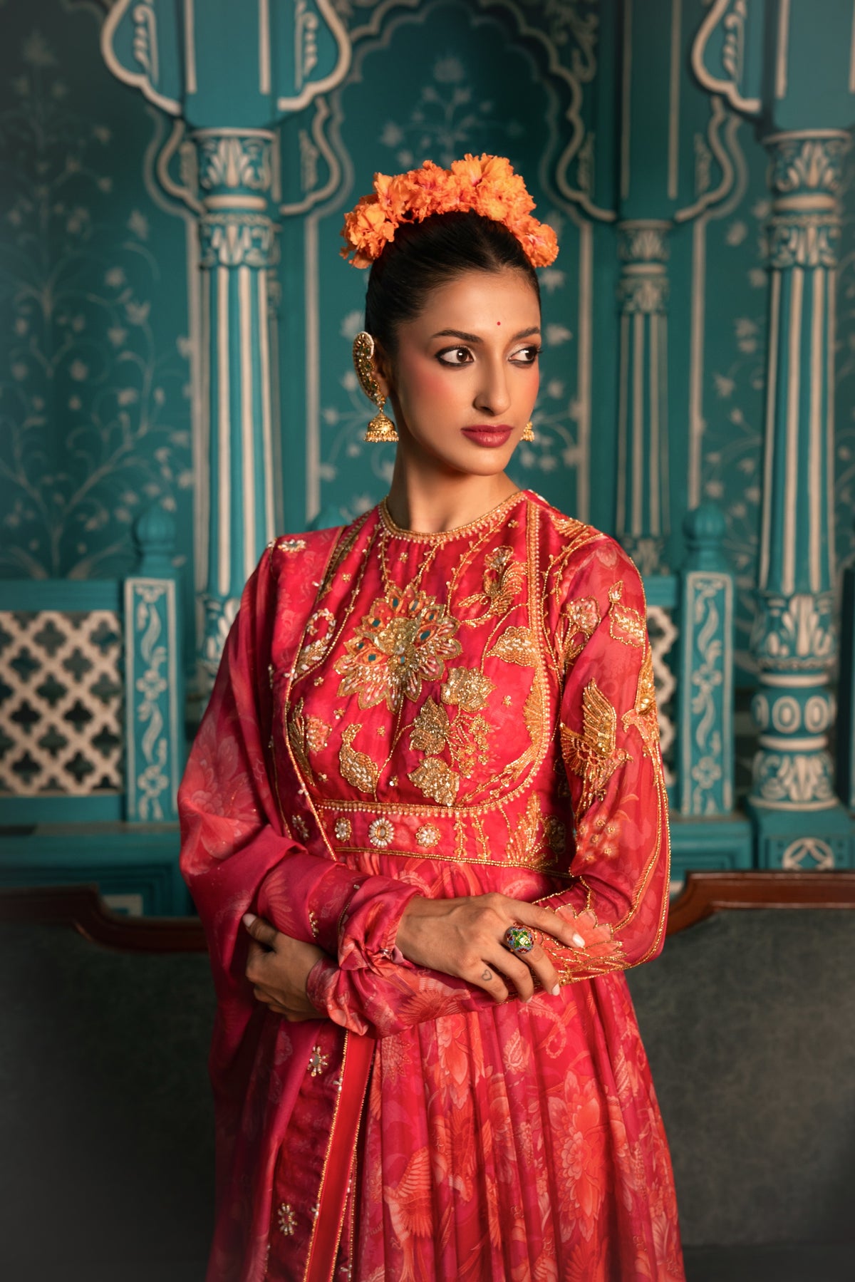 A woman wearing a traditional red and gold embroidered Indian outfit with an orange floral headpiece, standing in front of a teal patterned architectural backdrop.