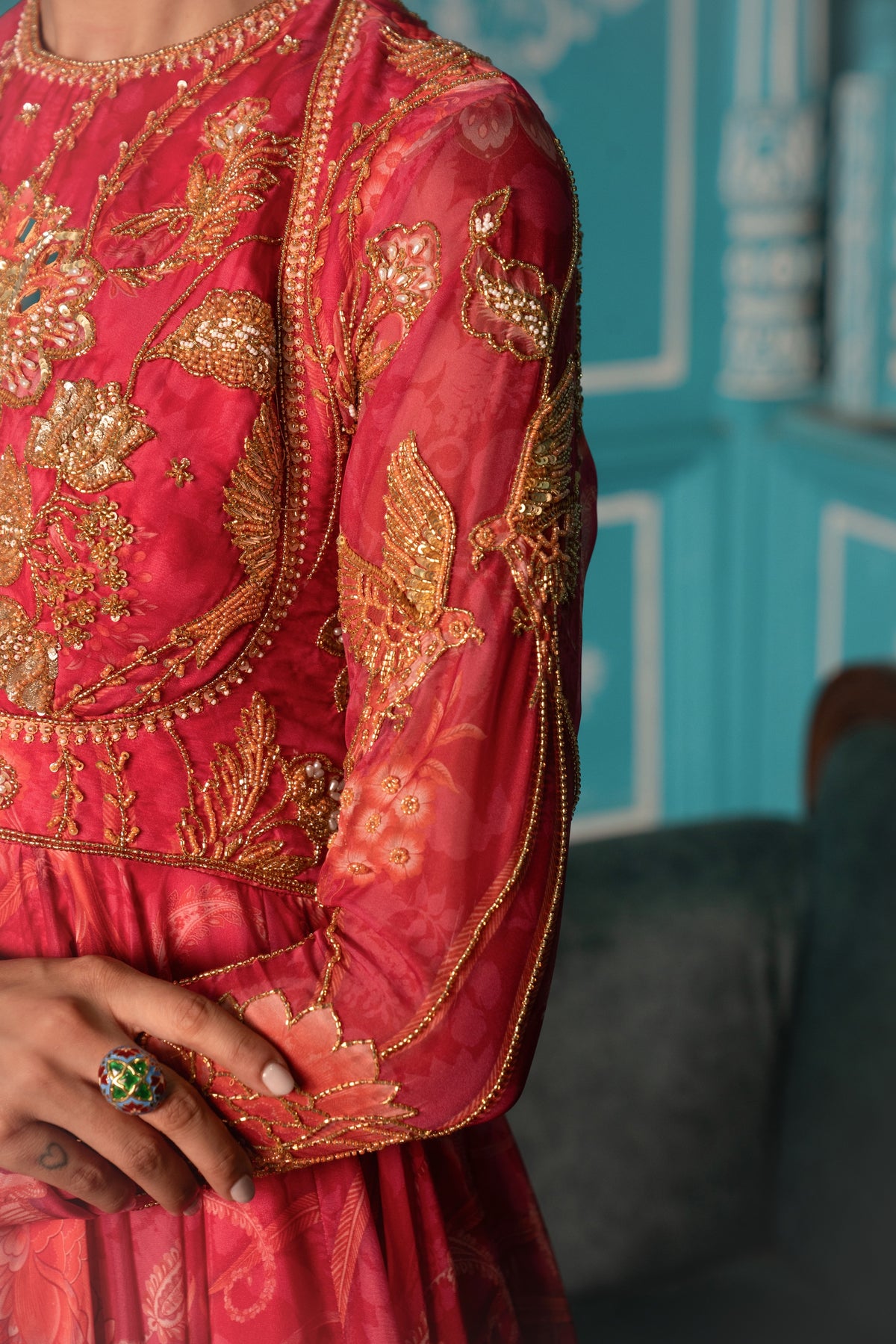 A close-up of a red and gold embroidered traditional garment with intricate metallic embroidery on sheer sleeves, worn by a model whose hand with a green ring is visible.