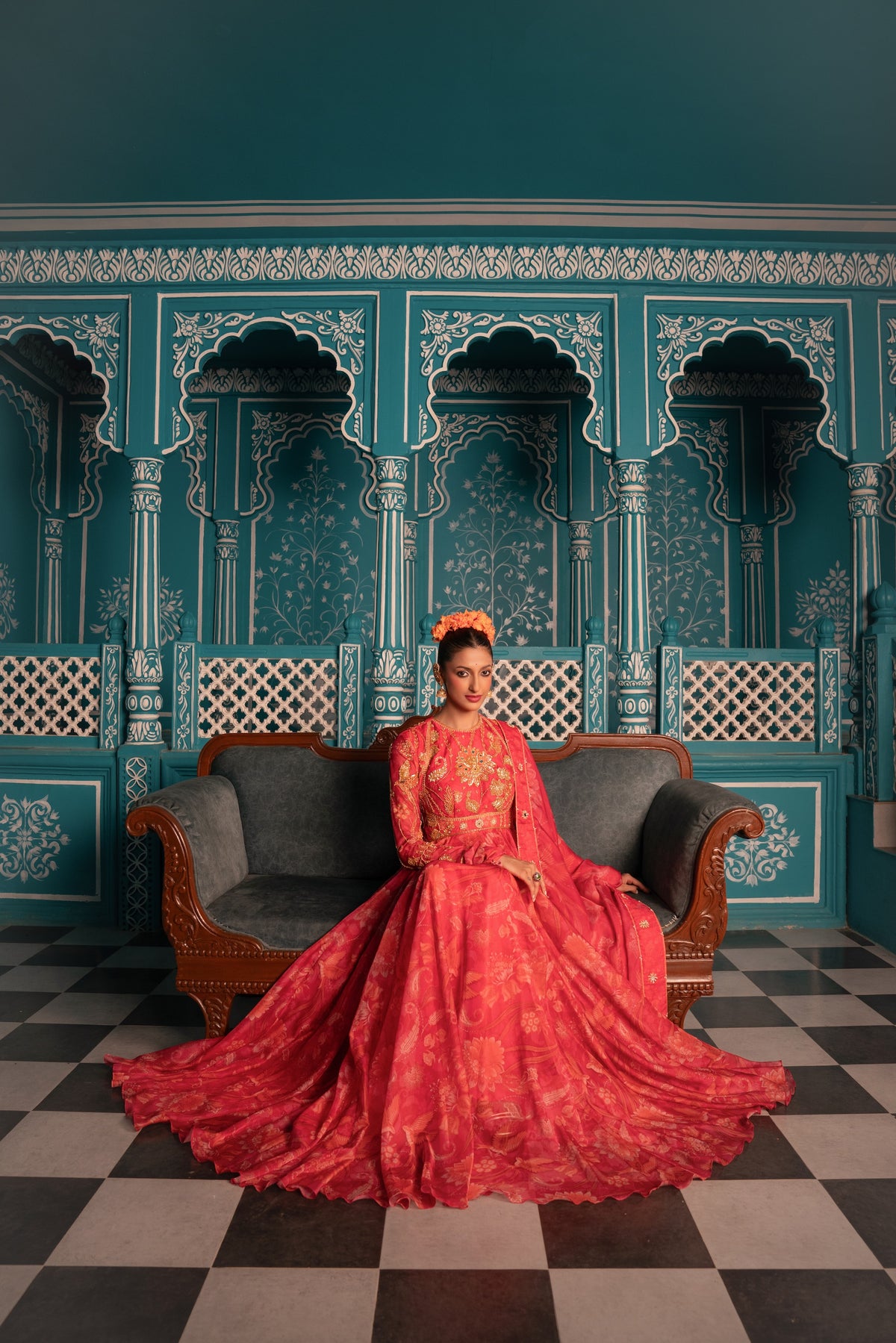 A woman wearing a vibrant red and pink floral lehenga choli sits on a dark gray sofa in front of a teal Moroccan-style wall with arches and a black-and-white checkered floor.