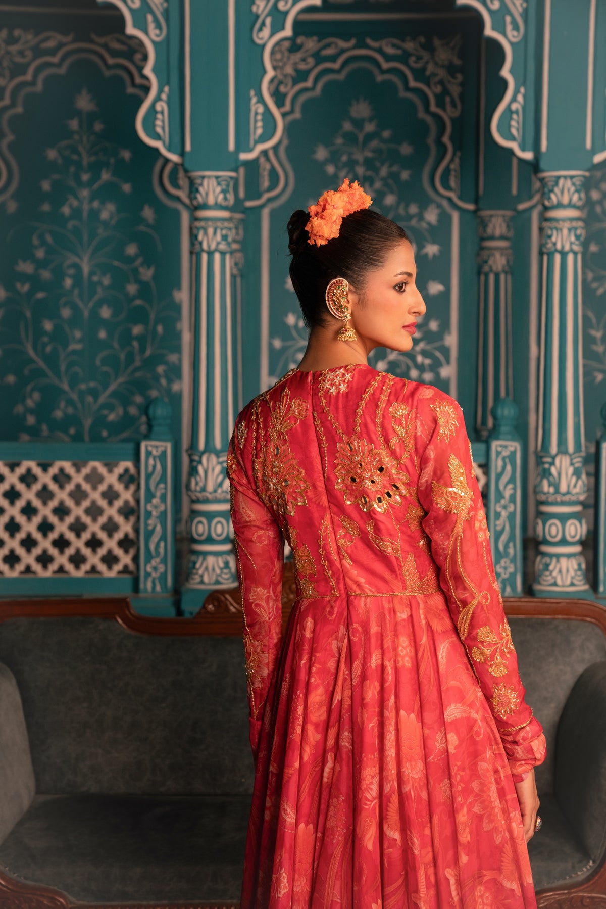 A woman wearing a red and gold embroidered traditional dress stands with her back turned, showcasing the detailed gold patterns on the back of the gown. She is posed in front of an ornate teal and white carved architectural wall.