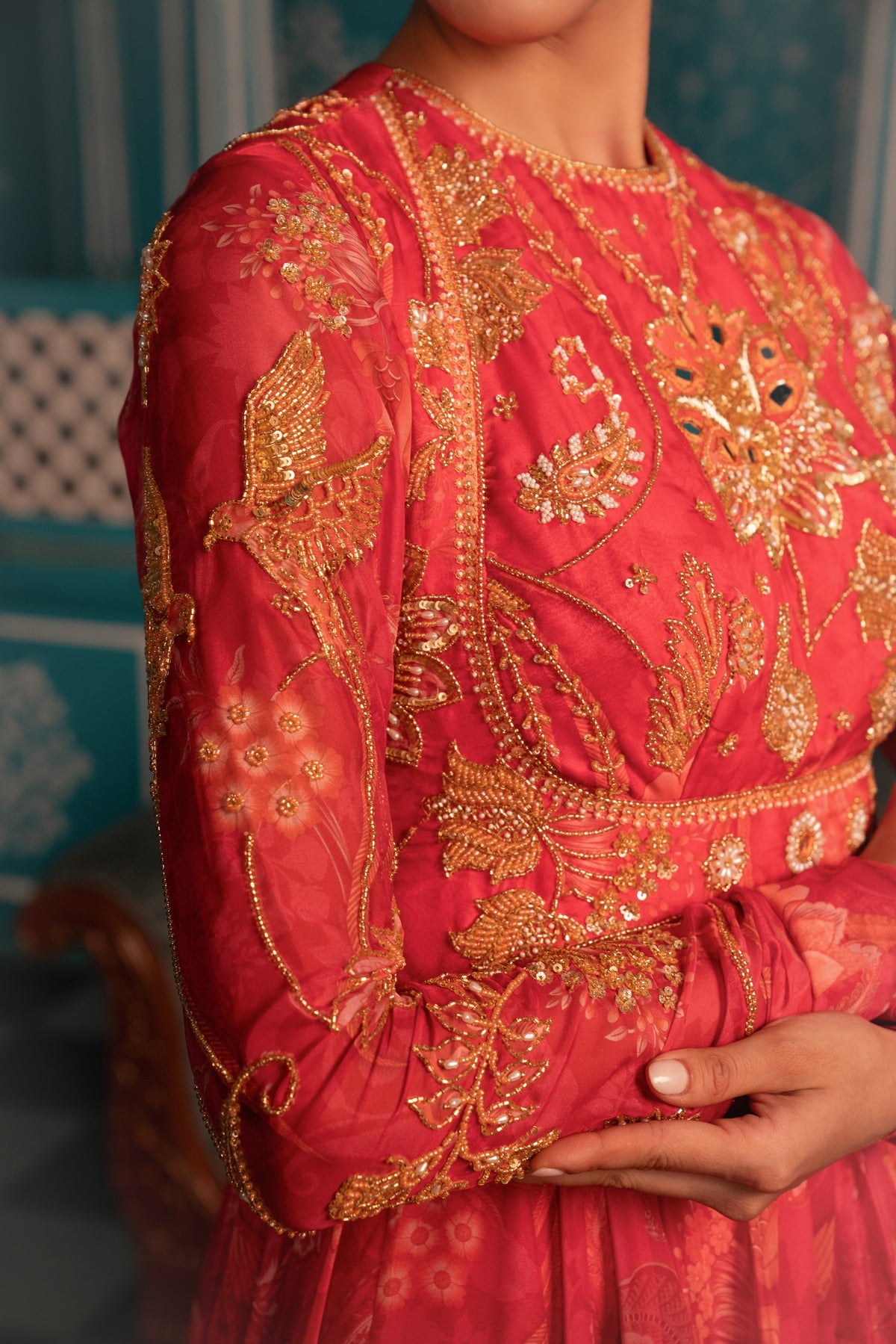 A close-up of a person wearing a bright coral-red embroidered dress with intricate gold metallic embroidery, beadwork, and floral motifs. The image shows the upper torso, one sleeve, and hands against a softly blurred teal interior background.