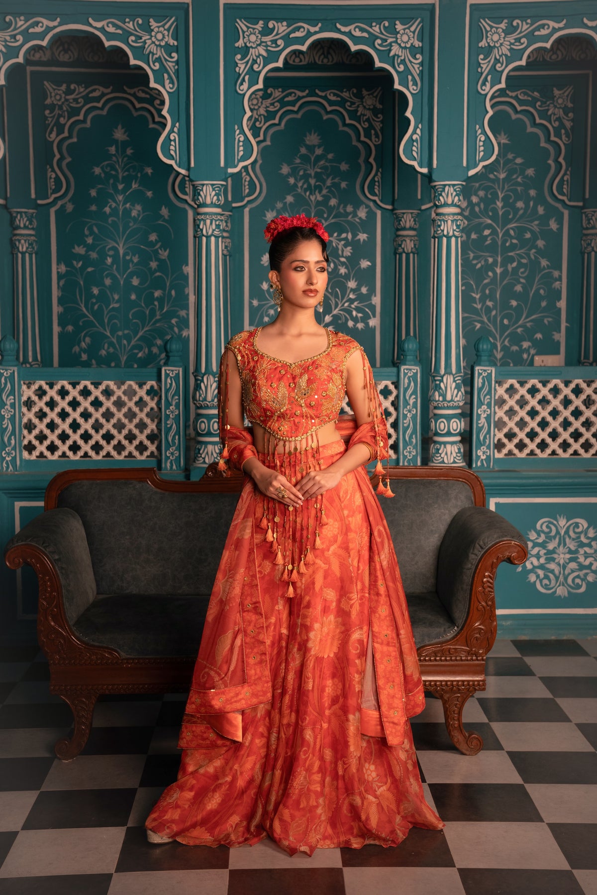 A woman stands in a traditional ornate room wearing a bright orange-red embroidered gown with sheer sleeves and a matching belt, posed in front of teal patterned arches and a dark sofa.