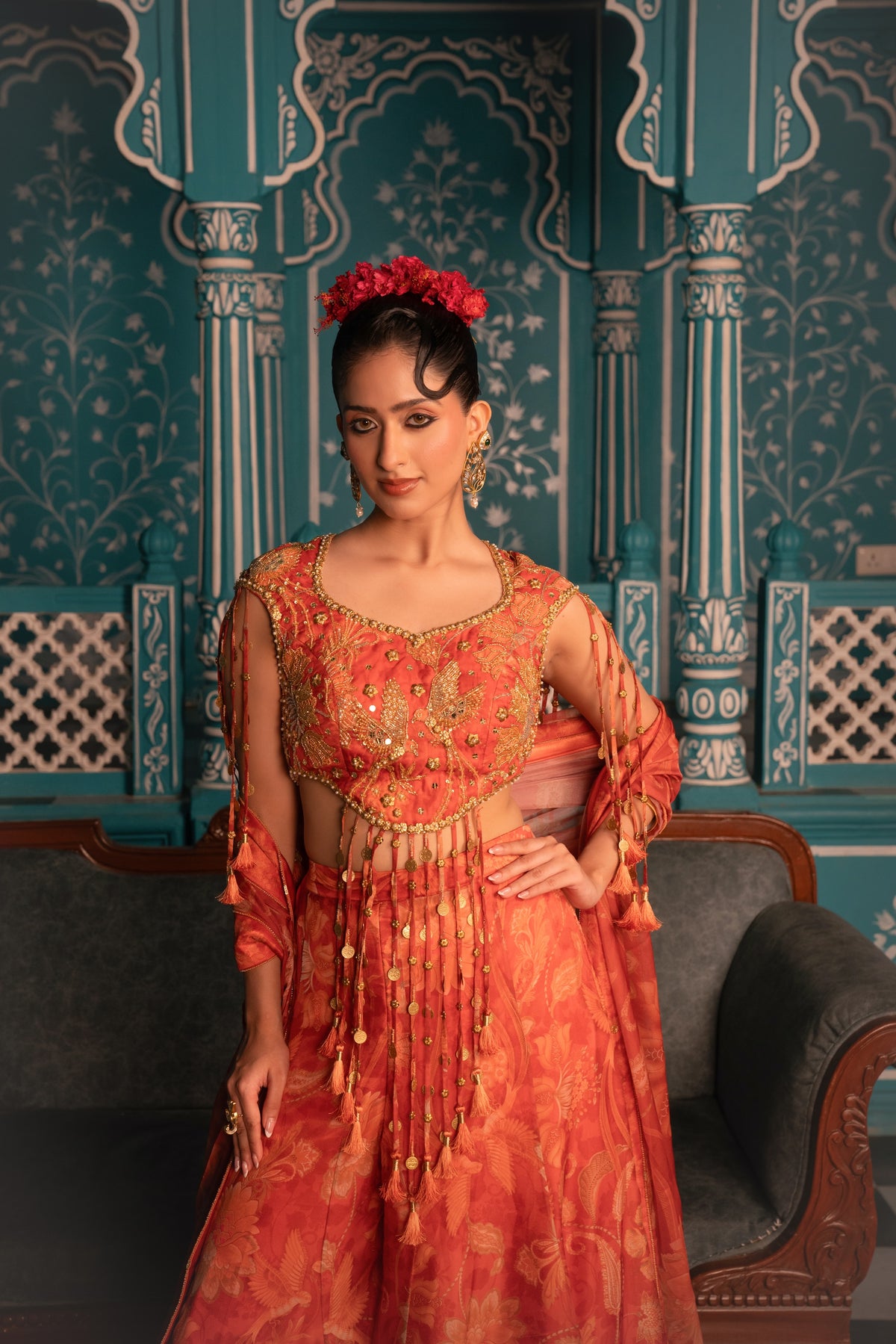 A woman models a traditional Indian salwar kameez outfit in a rich orange-red color with intricate gold embroidery, set against an ornate teal patterned wall.