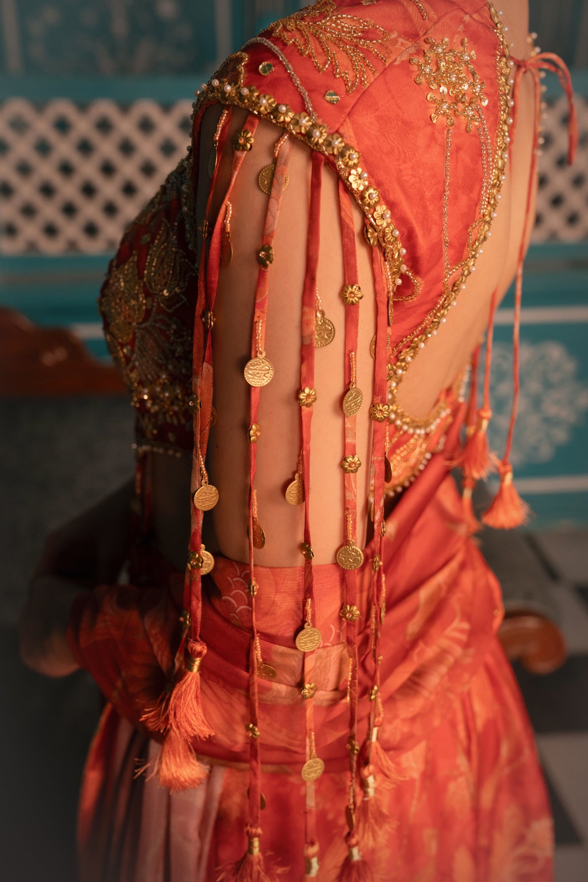 Close-up of a person wearing a richly decorated coral-orange traditional outfit with extensive gold embroidery, mirror work, and dangling coin garlands, photographed indoors against a teal patterned wall.