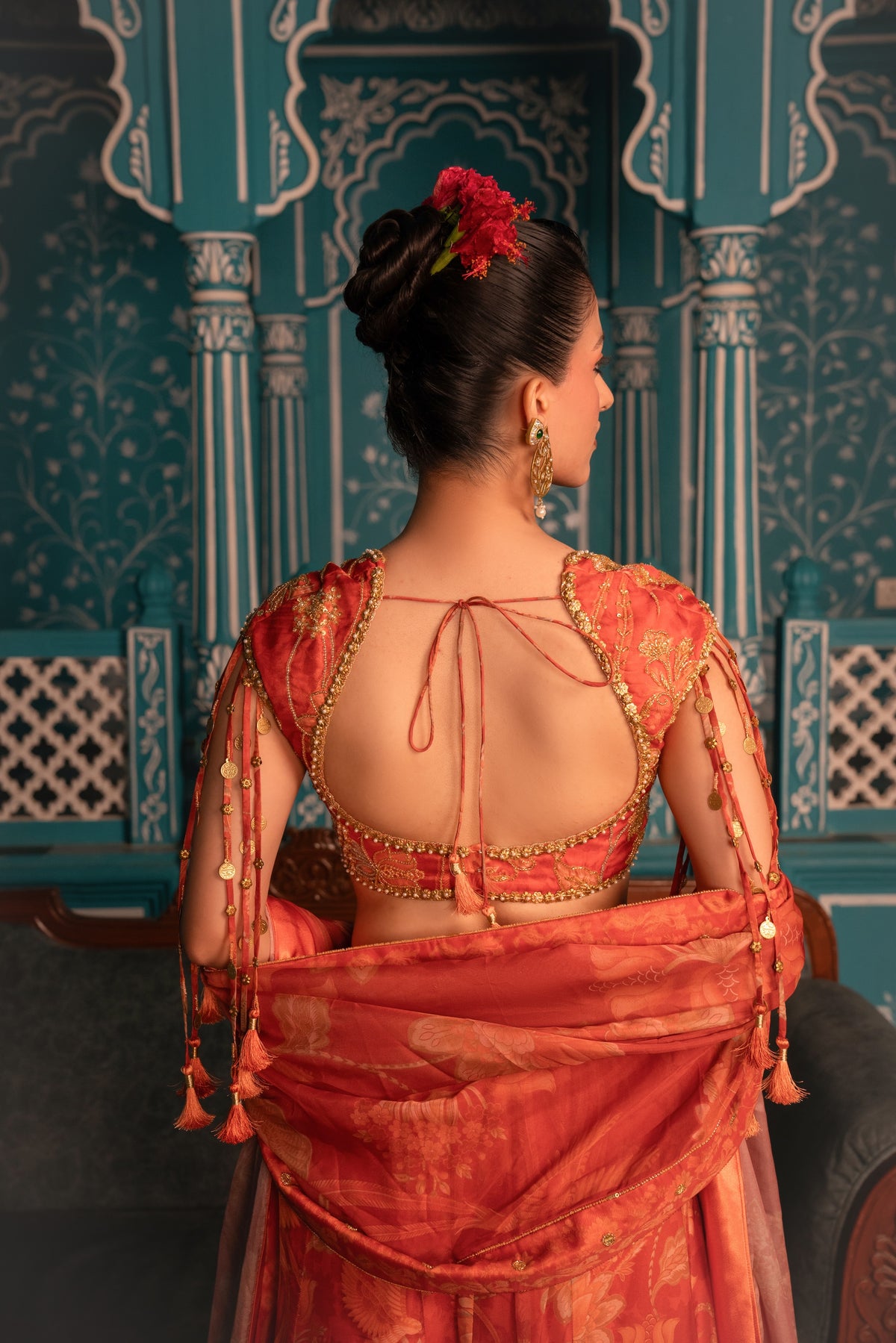 A woman viewed from behind wearing a traditional Indian saree with a red and orange floral print, featuring a gold-trimmed open back and sheer sleeves, set against an ornate teal architectural background.