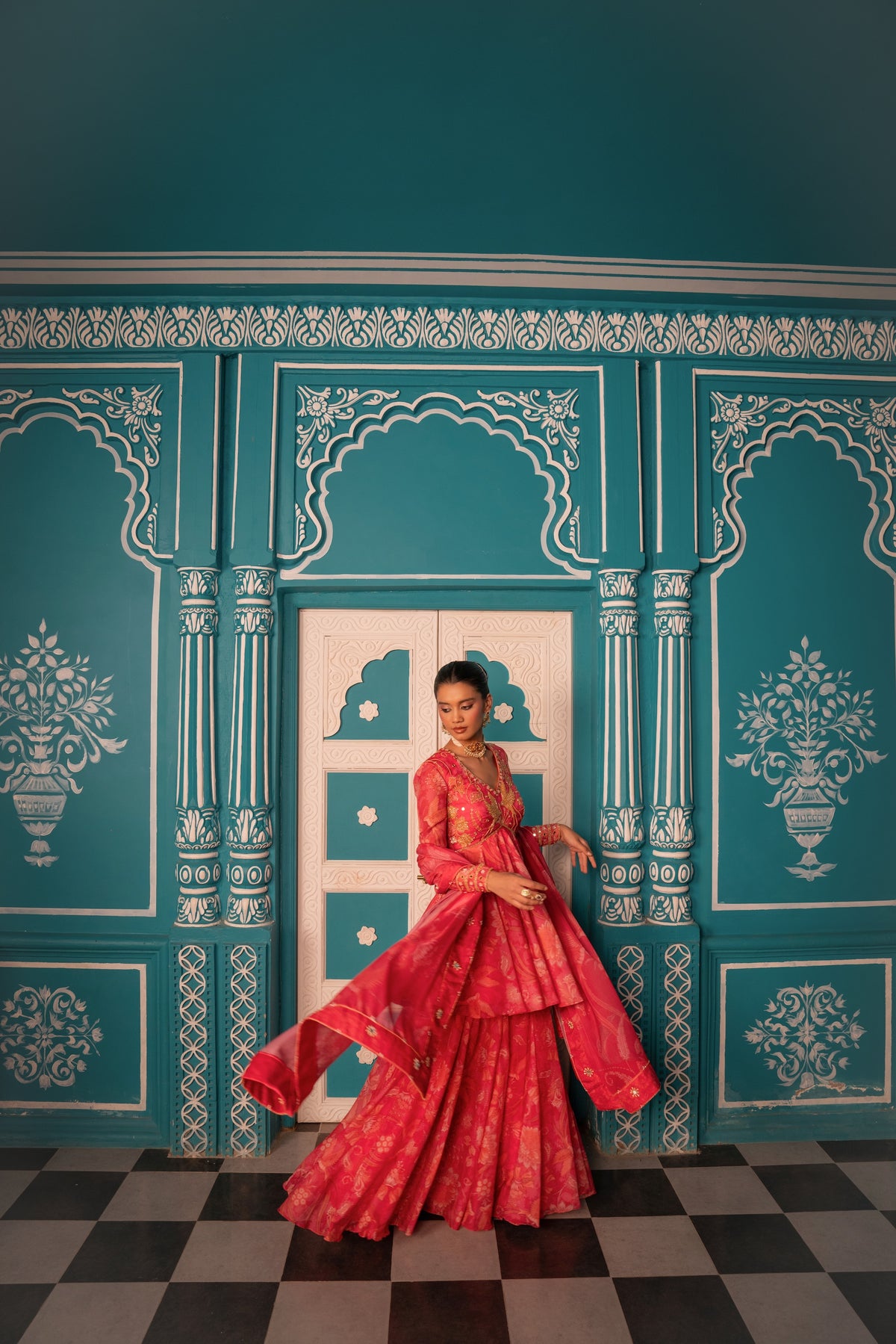 A woman wearing a vibrant red and pink embroidered traditional Indian outfit stands in front of an ornate teal and white paneled wall with a checkered floor.