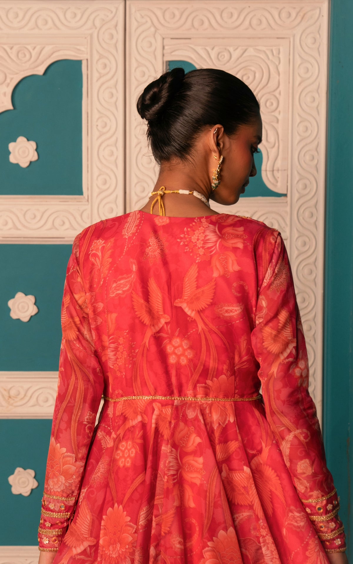 A woman shown from behind wearing a vibrant red and orange floral-patterned traditional dress with gold embroidery, standing in front of a decorative white and teal paneled door.