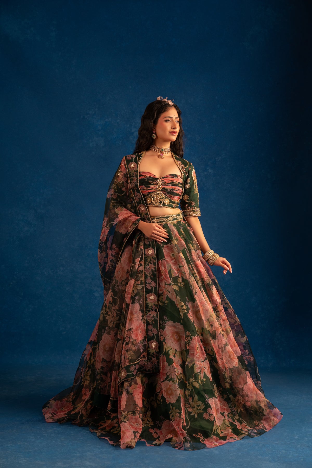 A woman models a traditional Indian lehenga choli set with a floral print, featuring a dark green base with pink and red flowers, set against a textured dark blue studio backdrop.