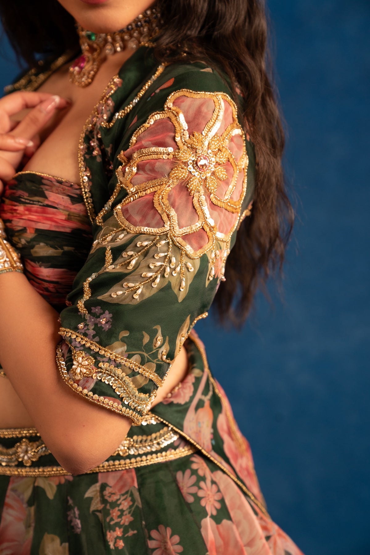 A close-up of a model wearing an ornate dark green embroidered traditional outfit with pink floral patterns and gold metallic embellishments, set against a solid blue background.