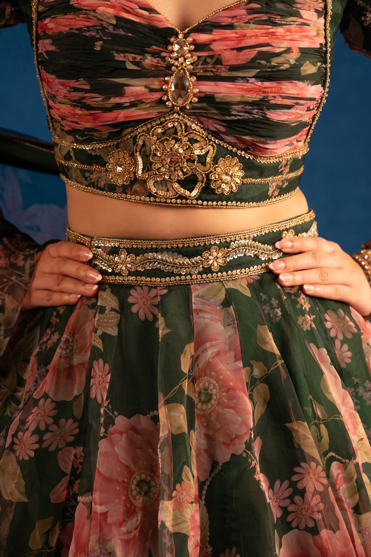 A close-up of a traditional Indian ethnic outfit featuring a dark green floral skirt with pink and beige flowers, a matching embroidered waistband, and a matching crop top with pink and green abstract patterns and gold embellishments. A person's hands are visible holding the waistband.