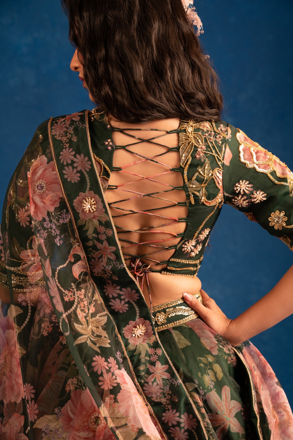 A woman shown from the back wearing a dark green floral printed outfit with a lace-up open back, pink and gold floral embroidery, and a matching sheer dupatta against a dark blue studio background.